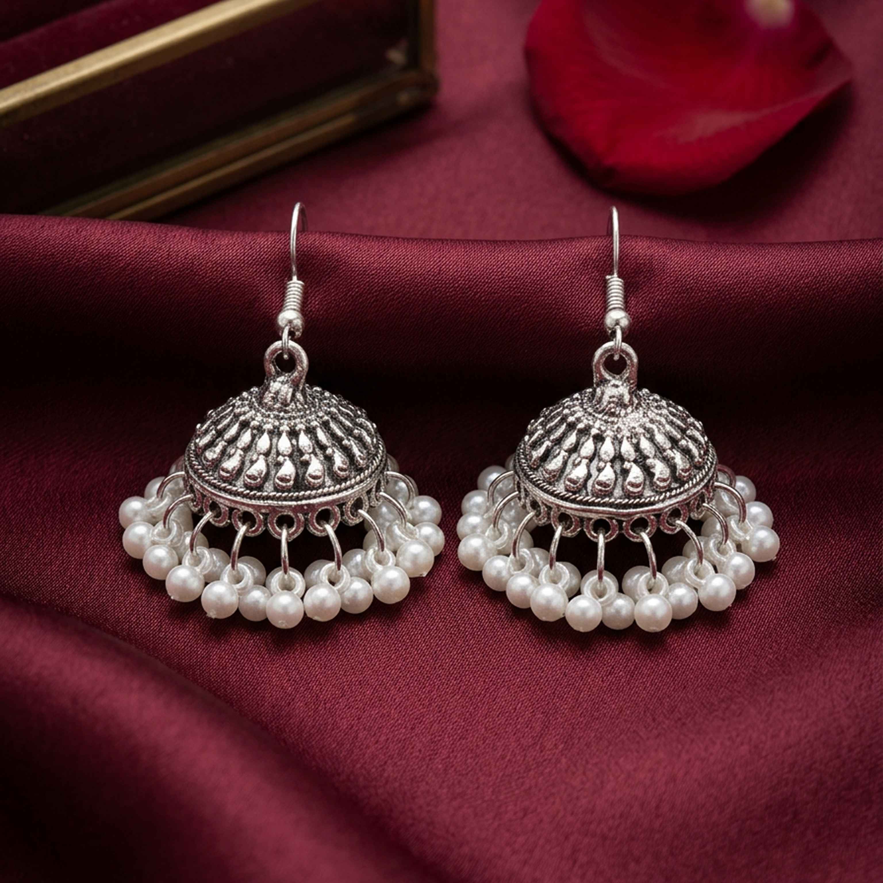 Pearl Jhumki earrings by Zaraya Store, two silver domed earrings with intricate designs and dangling white pearls on maroon fabric.