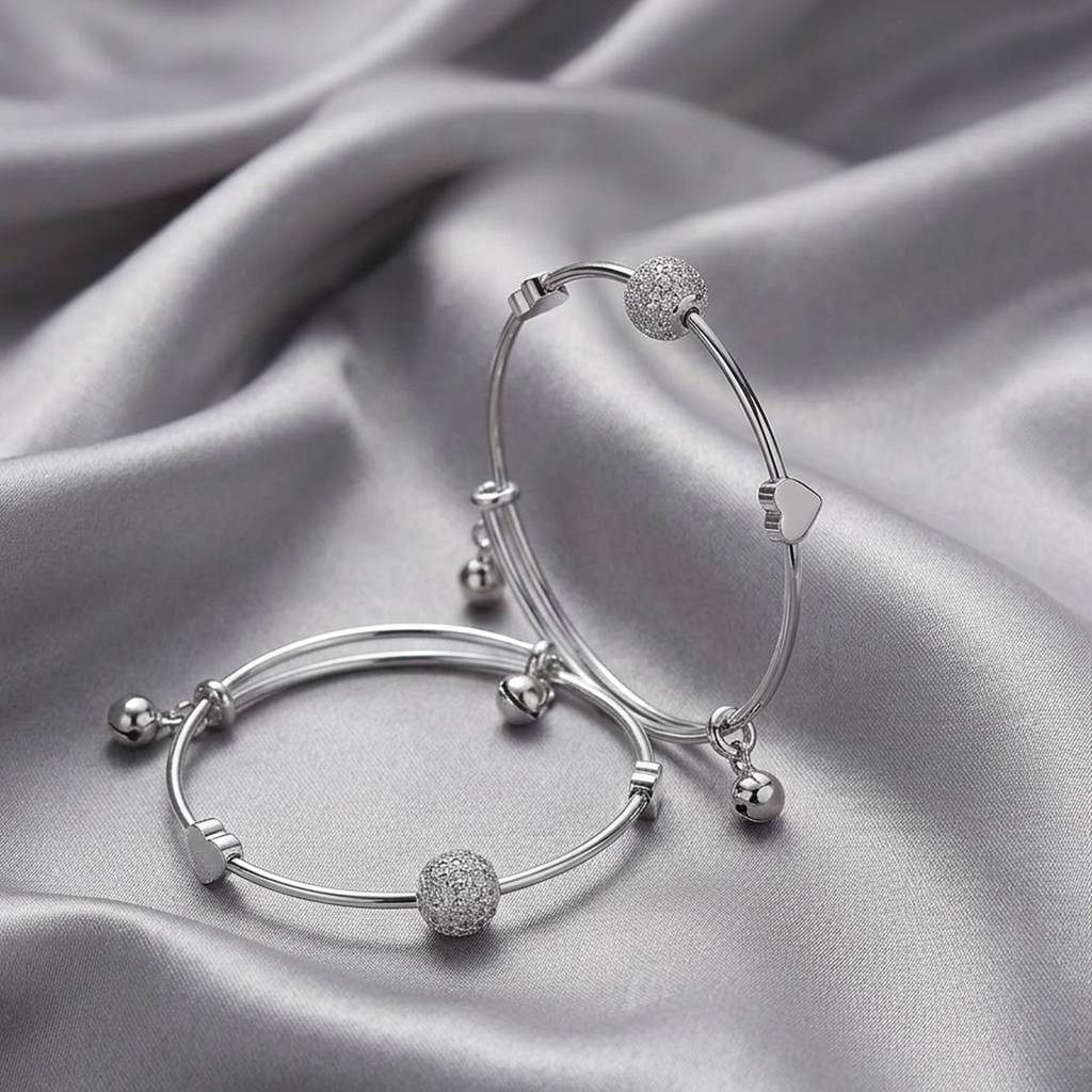 Set of two silver stainless steel adjustable bracelets with sparkling crystal orbs, heart charms, and small bells from Zaraya Store, lying on a flowing grey silk fabric.