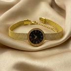 Gold mesh watch bracelet by Zaraya Store, with black dial, Roman numerals, Greek key bezel, on beige satin.