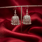 Antique silver jhumki earrings by Zaraya Store with intricate patterns and hanging metal beads, displayed against red fabric.