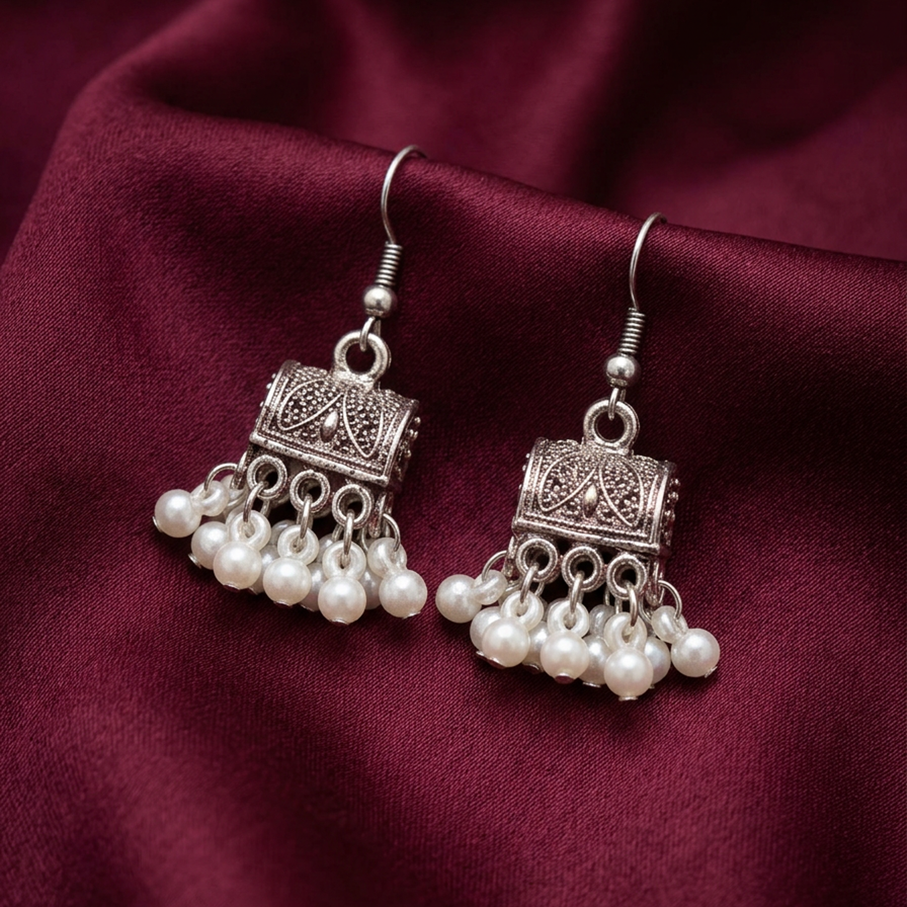 Antique white pearl jhumki earrings by Zaraya Store, with ornate silver-toned metal and hanging pearl drops on maroon fabric.