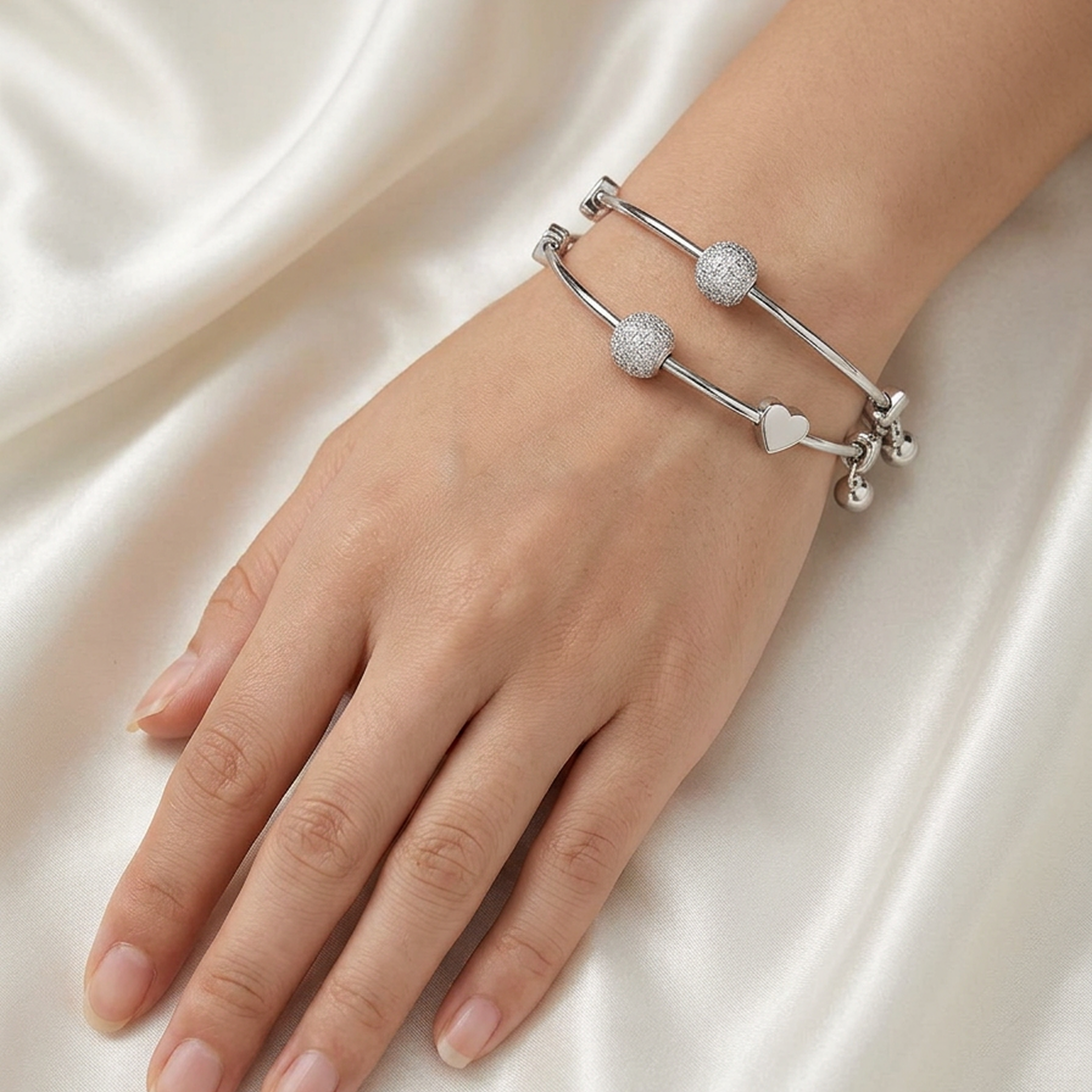 Silver stainless steel adjustable bracelets, a set of two, from Zaraya Store, with crystal orbs, a heart, and bells, on a hand against white satin.